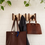 Wooden wall rack holding two brown handbags and a dark scarf against a light brick wall.