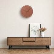 Round terracotta wall clock above a wooden media console with framed art and flowers.