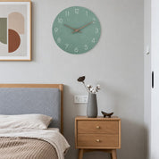 Sage green round wall clock with white numerals and copper hands above a wooden bedside table.