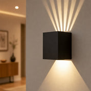 Black cube wall sconce casting warm geometric light beams on a neutral wall.