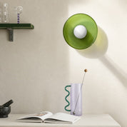 Green wall lamp above a lavender vase with wavy teal handle on a white tabletop.
