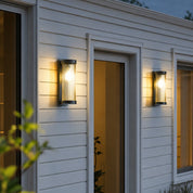 Two glowing cylindrical wall sconces mounted on a white house exterior at dusk.