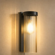 Lit ribbed glass wall sconce with black metal frame on a warm neutral wall.