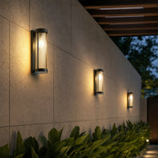 Three cylindrical ribbed wall lights glow warmly along a tiled outdoor garden wall.