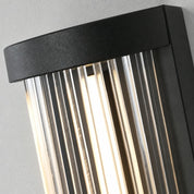 Black ribbed glass wall sconce glowing warmly against a light gray wall.