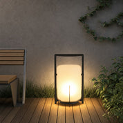 Glowing outdoor lantern with black frame on wooden decking beside plants.