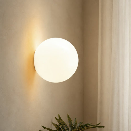 Glowing round wall light mounted above a leafy plant on a softly textured neutral wall.