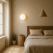 Glowing round wall sconce above a neutral linen bed in a warm minimalist bedroom.
