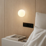 Glowing round wall lamp above a bedside table with a book and glasses next to white pillows.