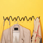 Black wavy wall rack holding a beige coat, chain strap, and cream scarf on a yellow wall.