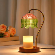Warm candle lamp with green and pink glass shade on a wooden base beside flowers.