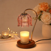 Pink glass flower-shade candle warmer lamp with a gold curved arm and wooden base beside a glowing candle.