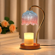 Lit candle warmer lamp with pink blue glass shade, gold arm, and wooden base beside peach roses.