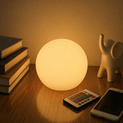 Glowing spherical lamp on a wooden table beside books, a remote, a phone, and a small elephant figurine.
