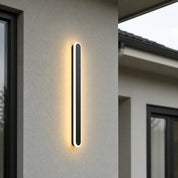 Vertical black outdoor wall light glowing warmly on a modern stucco exterior.
