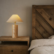 Wooden bedside table lamp with beige shade beside a rustic bed.
