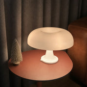 White glossy mushroom table lamp glowing on a coral side table beside a small decorative tree.