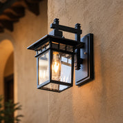 Black rectangular glass wall lantern with a glowing bulb mounted on a tan stucco wall.