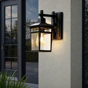 Black outdoor wall lantern with glass panels glowing on a light stucco exterior.