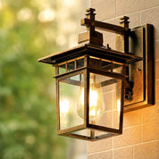 Bronze wall-mounted glass lantern with a glowing bulb on an outdoor brick wall.
