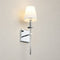 Chrome wall sconce with a glowing white fabric shade on a light neutral wall.