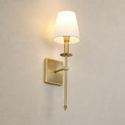 Brass wall sconce with a glowing cream shade on a plain light gray wall.