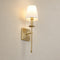 Brass wall sconce with a glowing cream shade on a plain light gray wall.