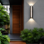 Vertical wall light glowing beside a wooden entry door among lush tropical plants.