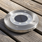 Round stainless steel solar deck light with LEDs and central panel on wooden planks.