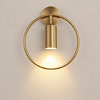 Brushed gold circular wall sconce with a glowing downward cylinder light on a pale wall.