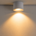 White cylindrical ceiling spotlight casts warm light on a plain beige wall.