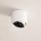 White cylindrical ceiling spotlight with a tilted black lens on a plain ceiling.