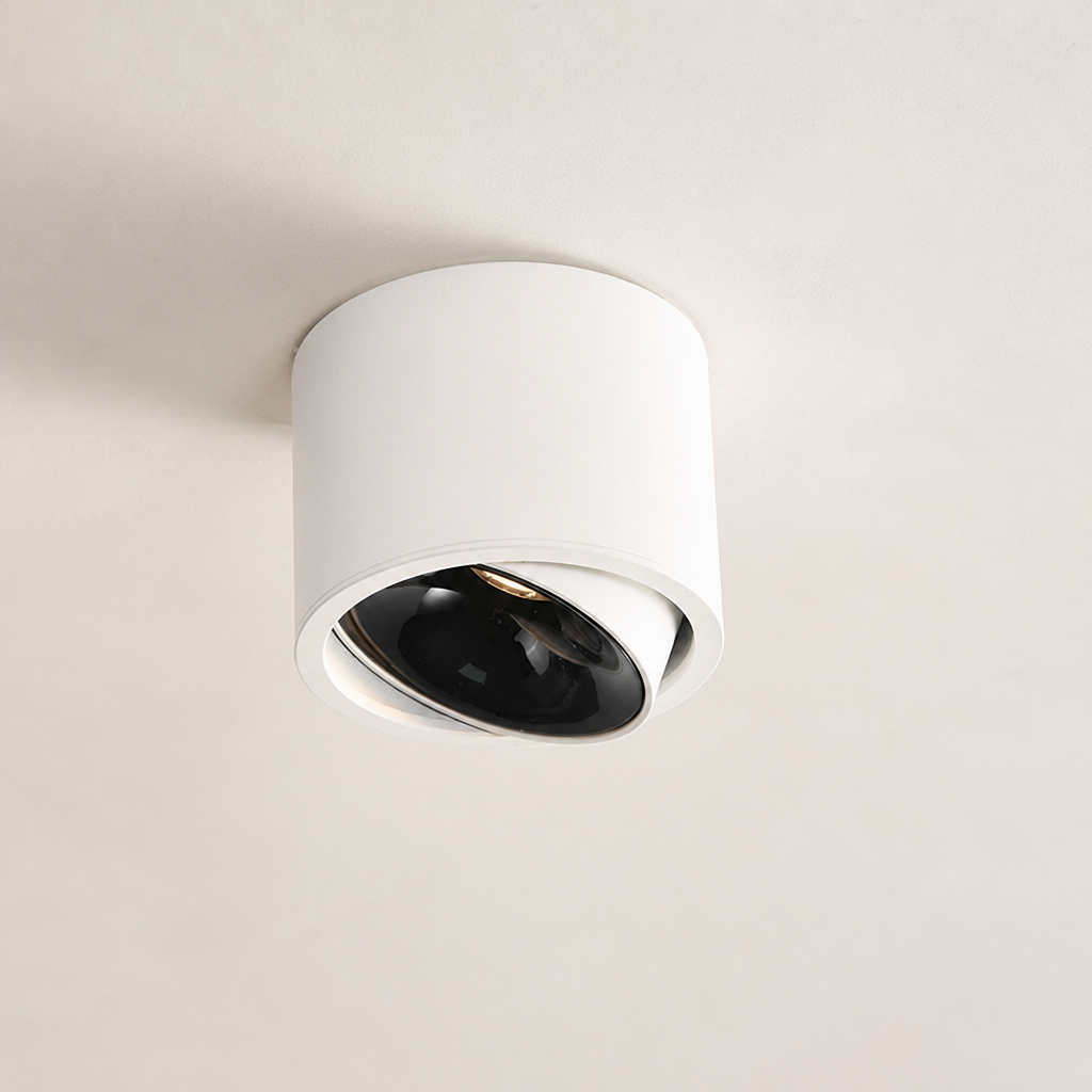 White cylindrical ceiling spotlight with a tilted black lens on a plain ceiling.