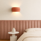 Terracotta ribbed wall sconce glowing above a pink upholstered bed headboard.