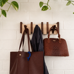 Wooden wall rack holding two brown handbags and a dark scarf against a light brick wall.