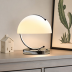 Glowing dome table lamp with chrome circular frame on a light wooden shelf.