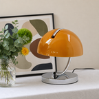 Glossy orange dome table lamp with chrome base on a white table beside flowers and framed art.