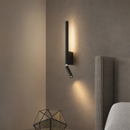 Black wall-mounted reading light beside a cushioned headboard with warm light on a taupe wall.