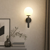 Black wall sconce with glowing white globe mounted above a console with books and a vase.