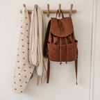 Brown leather backpack and patterned fabrics hanging from a wooden wall peg rail.