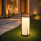 Glowing cylindrical outdoor lantern with a black frame on grass near a modern entryway.