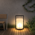 Glowing outdoor lantern with black frame on wooden decking beside plants.