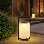 Glowing black framed outdoor lantern on a modern garden patio surrounded by plants.
