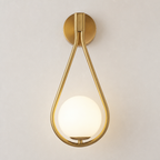 Brass teardrop wall sconce with a glowing white globe shade on a light neutral wall.