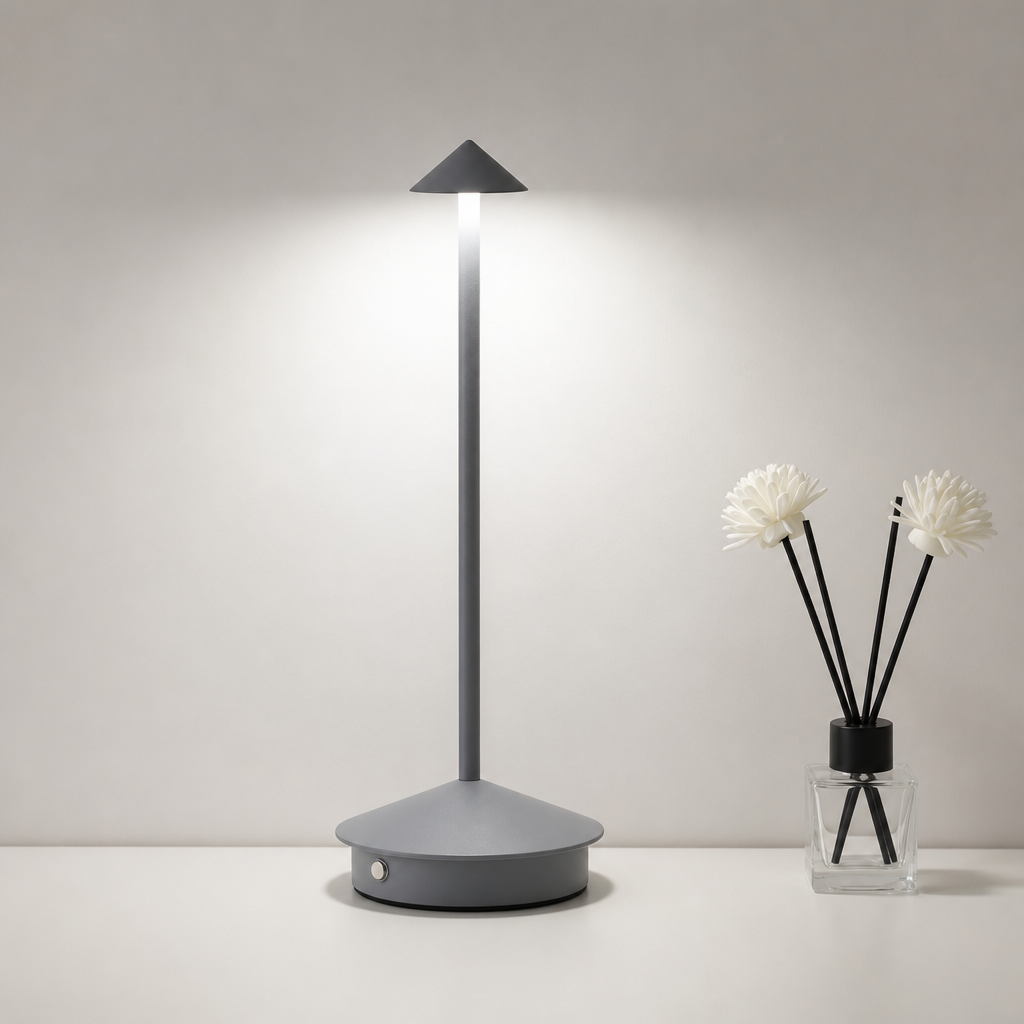 Gray cordless table lamp with a glowing cone shade beside a small reed diffuser.
