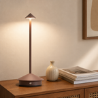 Brown slim cordless table lamp with conical shade and round base glowing on a wooden cabinet.